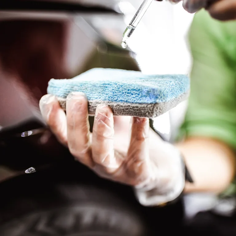 Close-up of a gloved hand holding a blue microfiber applicator pad while a drop of liquid ceramic coating falls from a pipette.