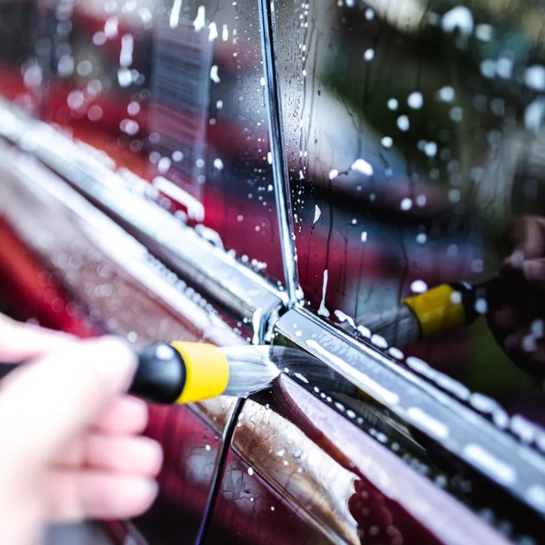 Close-up of a hand using a detailing brush to clean soapy residue from the trim of a red car door.