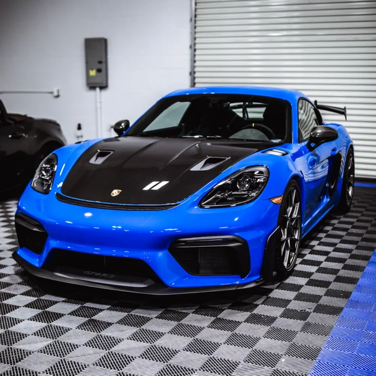 Bright blue Porsche GT4 RS with a carbon fiber hood parked indoors on a black and blue checkered floor.