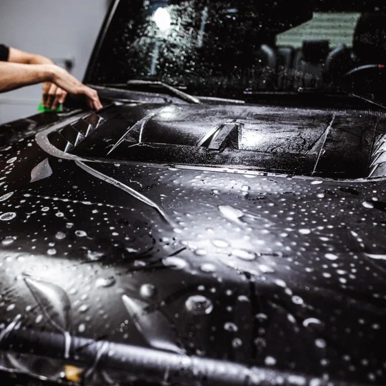 Close-up of a black vehicle hood with water droplets as a technician applies a protective film for paint protection.