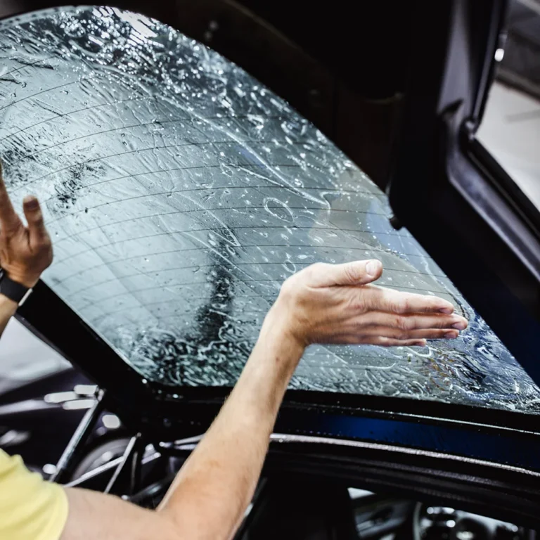 Person applying tint film to a car’s rear window, smoothing it with hands to remove bubbles.
