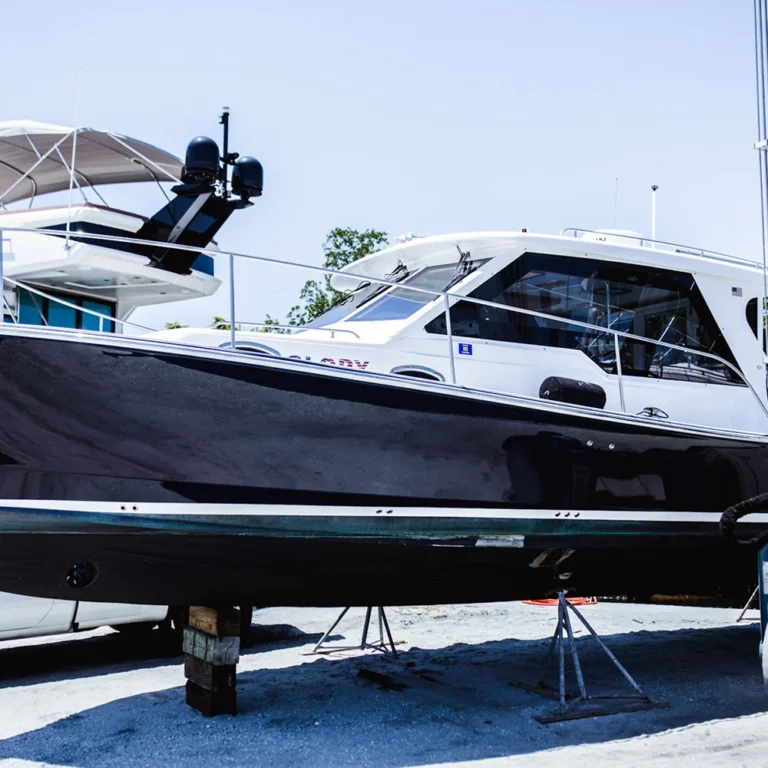 A black and white cabin cruiser named "Clarity" is propped up on supports in a boatyard, with neighboring yachts and a clear blue sky in the background.