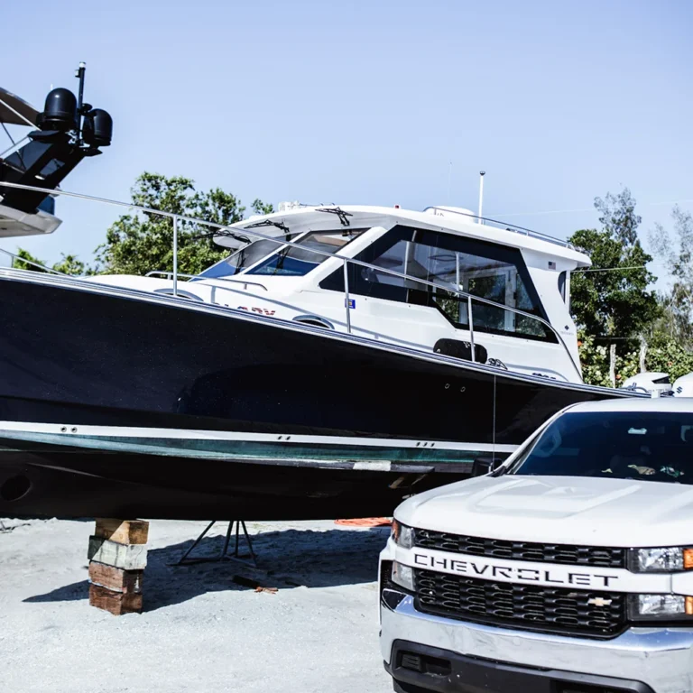 A black and white motorboat is elevated on wooden blocks for maintenance beside a white Chevrolet truck parked on a sandy lot, with trees and other boats in the background.
