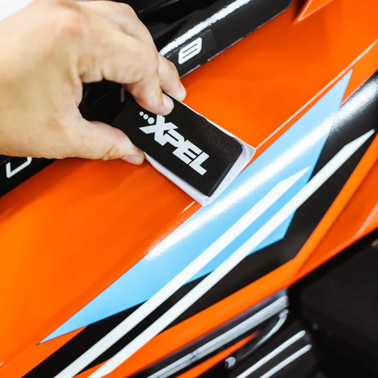 Hand using an XPEL-branded applicator tool to smooth protective film over an orange surface with blue, white, and black graphic accents. The film is being applied precisely for a seamless finish.