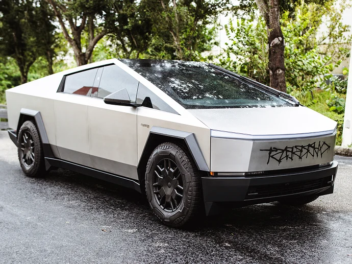 A silver Tesla Cybertruck with angular design is parked on a wet road, featuring a matte finish, large black wheels, and stylized graffiti-like text on the front.