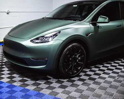 Matte green Tesla Model Y with black wheels parked indoors on a blue and gray checkered garage floor under bright lighting.