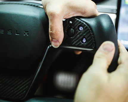 Close-up of hands gripping a Tesla yoke steering wheel, showing illuminated buttons for windshield wipers and voice control on a carbon fiber panel.