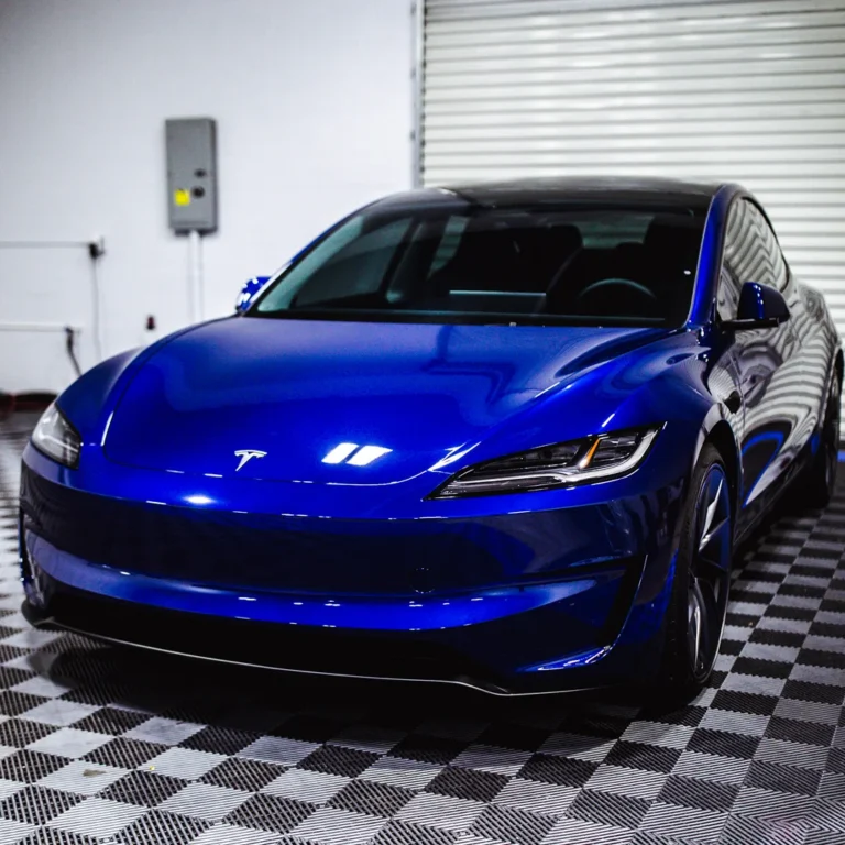 Shiny blue Tesla Model S parked indoors on a checkered floor, with reflections of overhead lights on its smooth hood and sleek body.