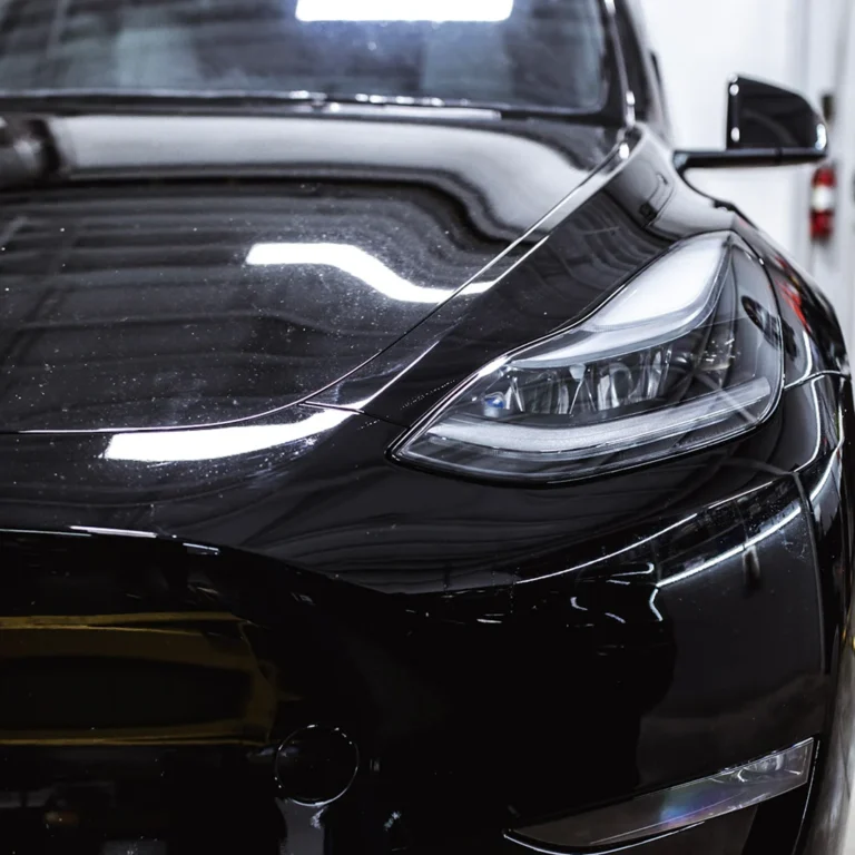 Close-up of a glossy black Tesla showing the front left headlight and part of the hood, reflecting ceiling lights and surrounding structures.