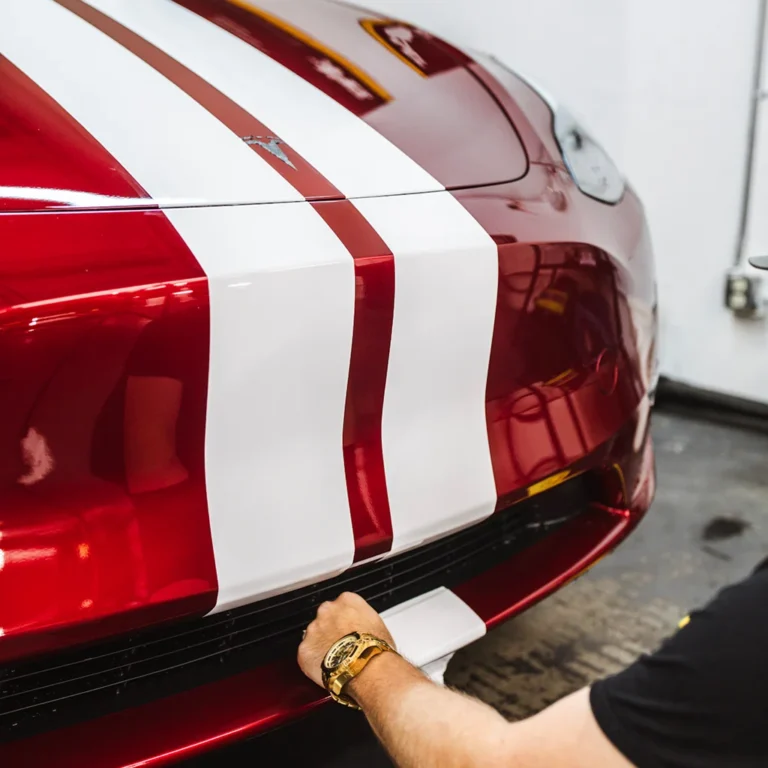 Close-up of a red Tesla with bold white racing stripes being detailed by a person wearing a gold watch, focusing on the front bumper area.