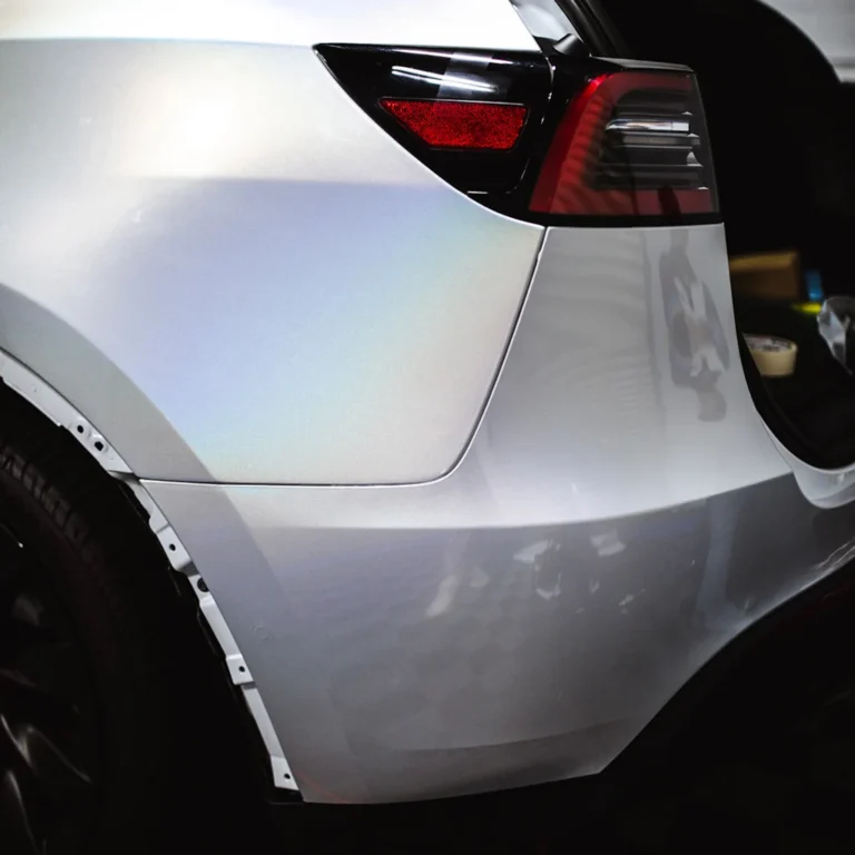 Close-up of the rear quarter panel of a white Tesla with an iridescent finish, showing the taillight, open trunk, and partially removed trim near the wheel well.