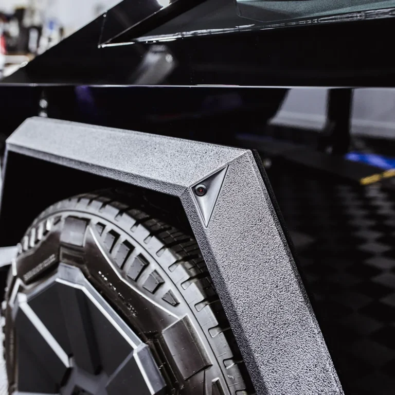 Close-up of a Tesla Cybertruck’s rugged rear fender with textured coating, angular wheel arch design, and embedded side camera above the large all-terrain tire.