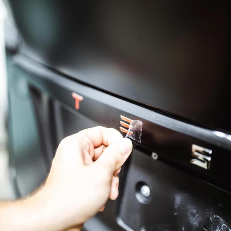 Close-up of a hand peeling a protective film from custom red and chrome badging on the matte black tailgate of a Tesla Cybertruck.