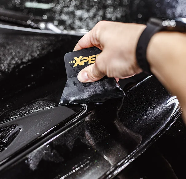 Close-up of a person applying XPEL paint protection film to a black car hood using a squeegee tool.