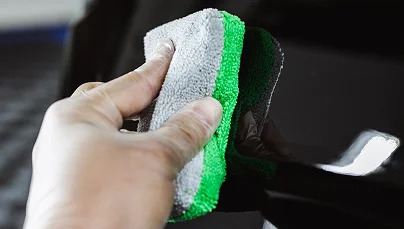 Close-up of a hand using a gray and green microfiber cloth to clean the surface of a black vehicle.