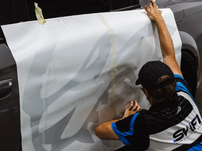 Technician applying a large vinyl graphic to the side of a gray vehicle, smoothing the material for proper adhesion.