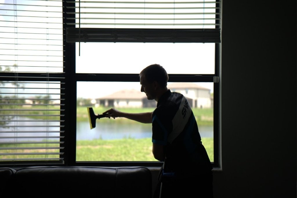 A technician from Southwest Florida Tinting uses a squeegee to finish installing residential window film inside a Fort Myers home, with a lake and neighborhood visible through the window. The scene reinforces why Residential Window Tinting is Worth the investment for comfort, efficiency, and UV protection.