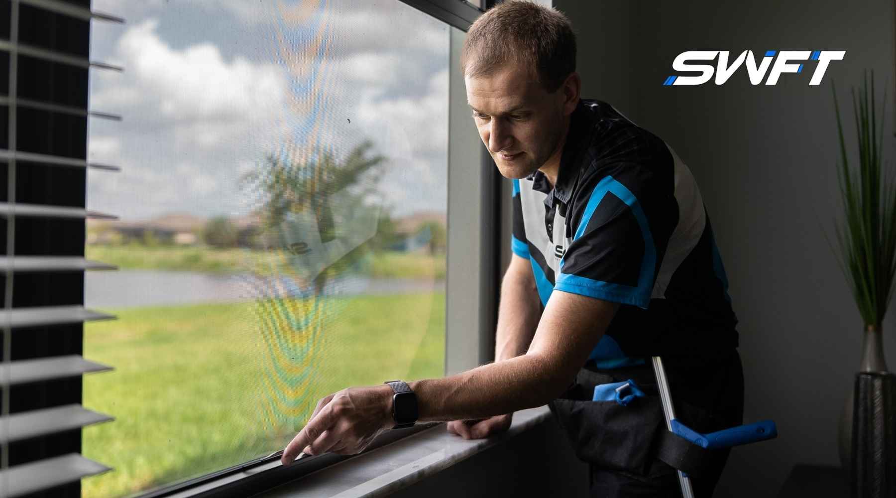 A technician from Southwest Florida Tinting installs residential window film in a Fort Myers home, carefully applying the tint to a large window overlooking a green lawn and lake. The image supports the topic of Residential Window Tinting Cost by visually illustrating the professional service in progress.