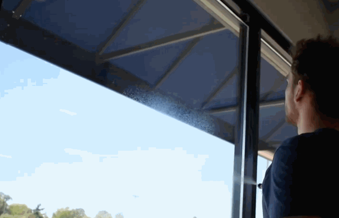 A person sprays a clear liquid onto a large indoor window as part of a commercial window tinting process. The outside view shows a sunny day, indicating glare and heat exposure.