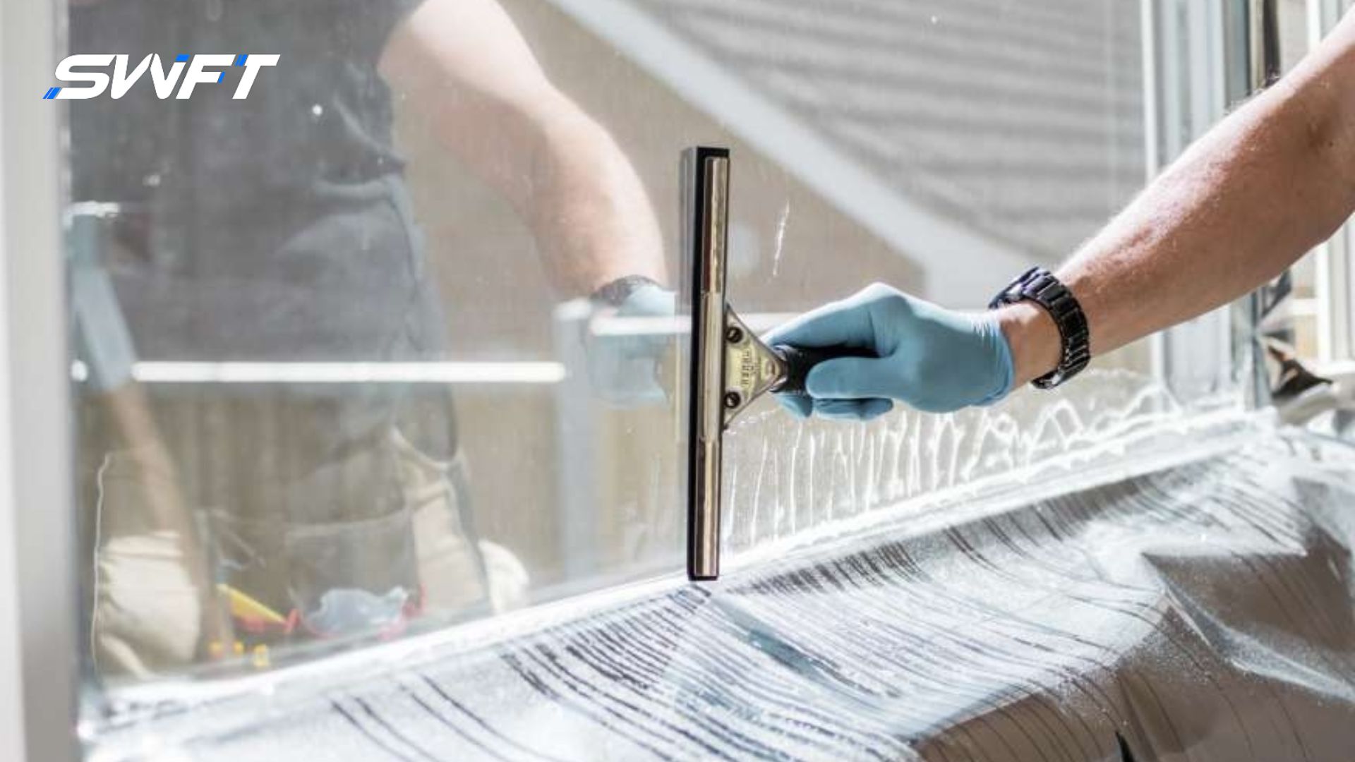 A worker wearing gloves uses a squeegee to clean a window before applying commercial window tint film, with the SWFT logo in the corner.