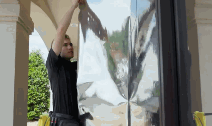 A technician stretches a shiny, reflective window film over a commercial glass door, preparing it for installation.