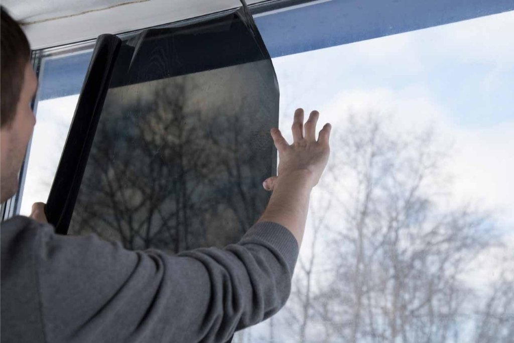 A person installs a dark residential window tint film on a large home window, carefully aligning it to cover the glass. This visual supports the topic of Typical Residential Window Tinting Cost by showing part of the hands-on process involved in improving energy efficiency and comfort.