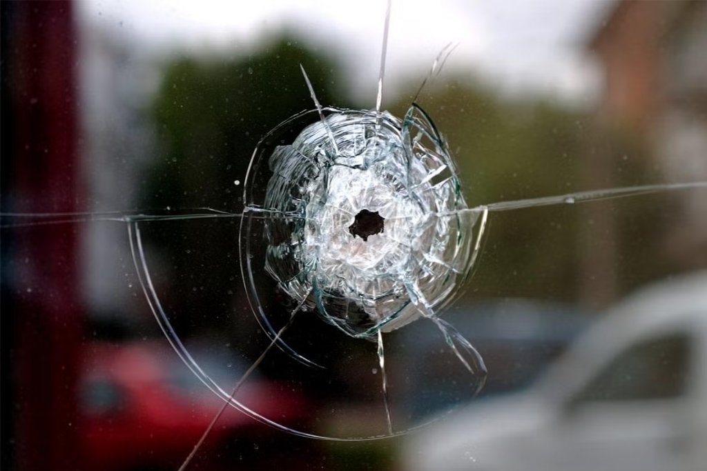 A close-up of a glass window with a bullet hole and spiderweb cracks shows the damage from a high-velocity impact, emphasizing that while security window film can resist shattering, it is not bulletproof. This image illustrates the key limitation of what Security Window Film Doesn't Do in extreme threat scenarios.