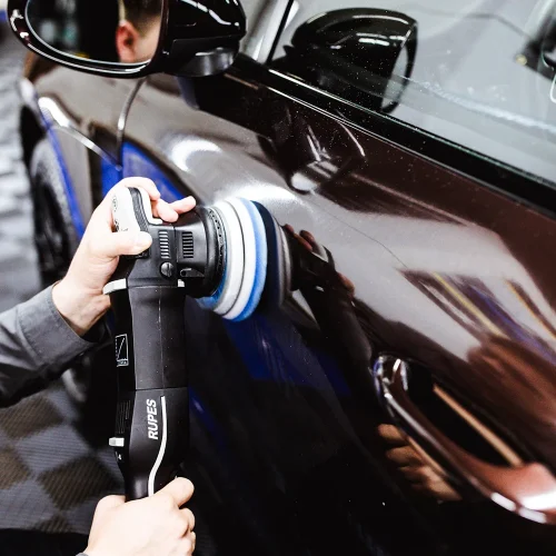 Person using a Rupes polishing machine to buff the side of a black car, enhancing the paint’s shine and removing imperfections. Reflections of the surroundings are visible on the glossy car surface.