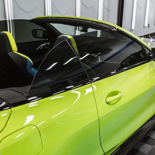 Close-up side view of a vibrant lime green convertible sports car with black trim and tinted windows. The car’s sleek design and glossy paint finish reflect the surrounding lights in the garage.