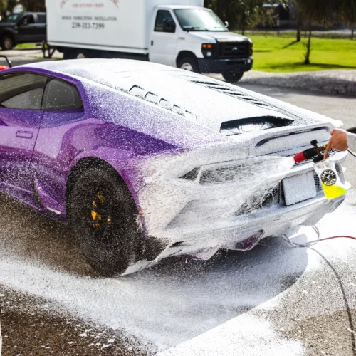 A purple sports car is being coated in thick white foam during an outdoor wash, with soap covering most of its rear and side panels. A person uses a foam cannon attachment to apply the suds under bright daylight.