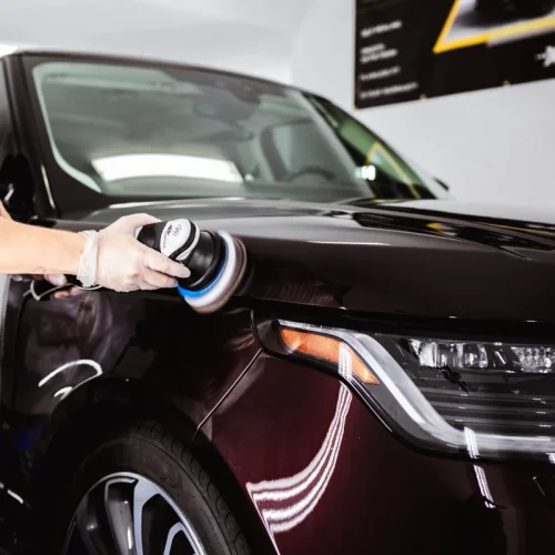 Detailer wearing gloves uses a dual-action polisher to buff the hood of a dark maroon luxury SUV, enhancing the paint’s gloss under bright shop lighting. The vehicle's front headlight and alloy wheel are visible, reflecting the clean, polished finish.