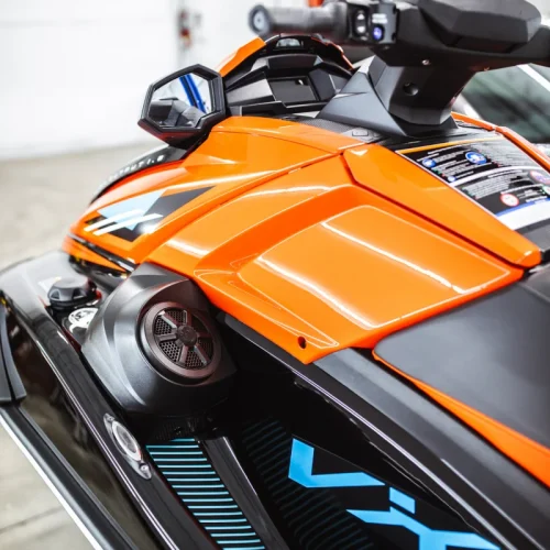 Close-up of an orange and black Yamaha VX Cruiser personal watercraft, showing the handlebar controls, side mirror, built-in speaker, and sleek body graphics. The jet ski is positioned indoors under bright lighting.