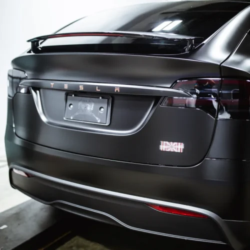Rear view of a matte black Tesla with tinted taillights and a subtle spoiler, parked indoors on a lift. The license plate area is empty, highlighting the sleek custom finish.