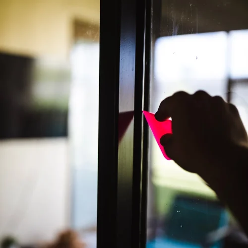 Person using a small pink squeegee to apply or smooth window film on a glass door, ensuring a clean and bubble-free finish. The scene is softly lit with a blurred background indoors.