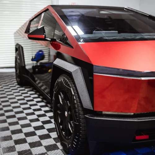 Tesla Cybertruck with a red and black two-tone finish parked indoors on a checkered garage floor. The angular front design and rugged wheel arches highlight its futuristic styling.