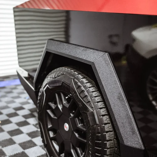 Close-up of a rugged black wheel and tire on a Tesla Cybertruck, showing its angular fender design. The background features a checkered garage floor and a partially visible white vehicle.